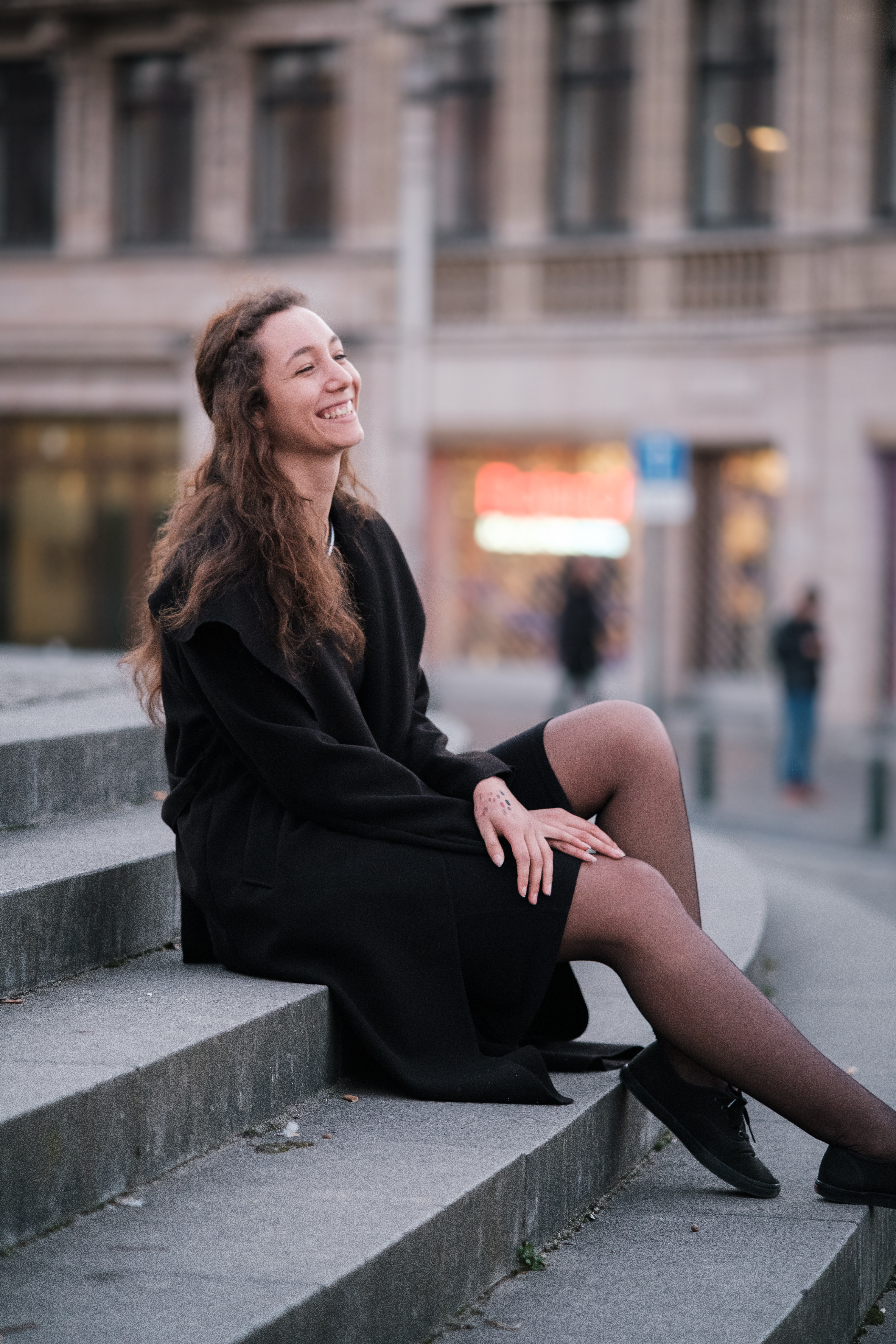 White/latina woman with long brown hair dressed in black, 
													   sitting outside on stairs, smiling 